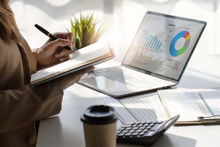 An individual holds a notebook, poised to write with a pen, with a calculator, clipboard and laptop open to charts and graphics is on the table in front of them